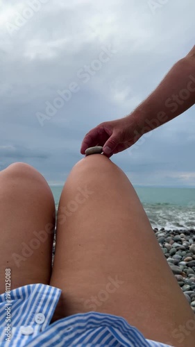 Vertical 4K video. 60 fps. Male hand builds small rock tower, stone pyramid on female leg knee at sun day. Concept of rest on pebble beach, summer vacation, ocean shore zen, tourism, travel, balance