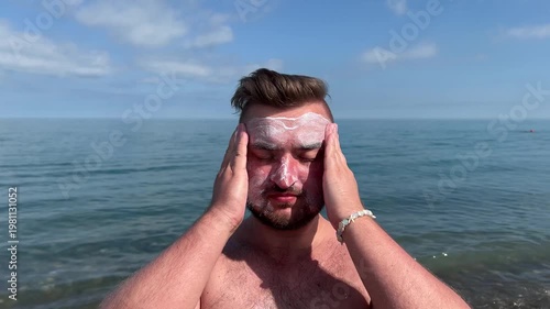 Travel 4K video. 60 fps. Young adult tanned white fat naked man with stubble applies milk balm, lotion, sunscreen cream to face on sea beach, ocean shore. Blue sky. Tourism, summer vacation, skin care