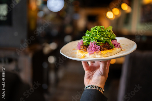 A waiter holds a white plate with a gourmet open-faced lamb burger topped with pickled red onions and fresh greens in a dimly lit, upscale restaurant or bar setting.