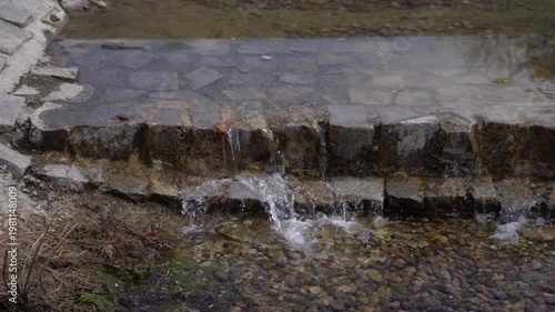 Water flows over a small stone ledge forming a miniature waterfall in a shallow pond