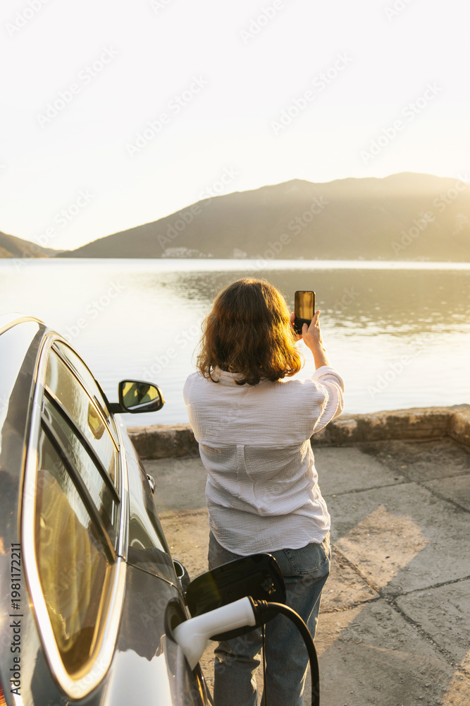 Fototapeta premium Woman with smartphone next to charging electric car on the seacost during summer vacation