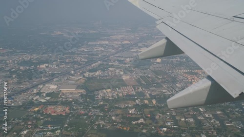View from airplane window flying over Bangkok city with wing visible during flight above urban landscape. Aerial travel concept