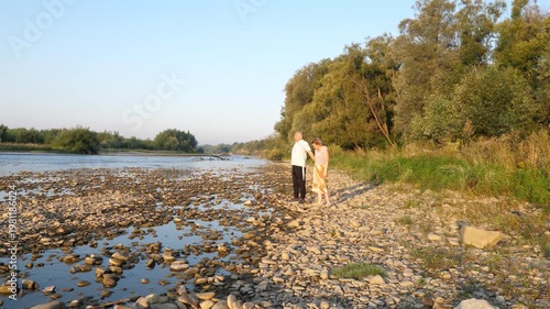 A man and a woman stand on a rocky riverbank watching the water under soft evening light