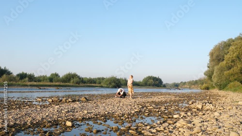 A man and a woman stand on a rocky riverbank watching the water under soft evening light