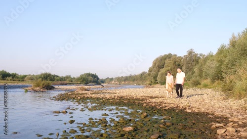 A man and a woman stand on a rocky riverbank watching the water under soft evening light