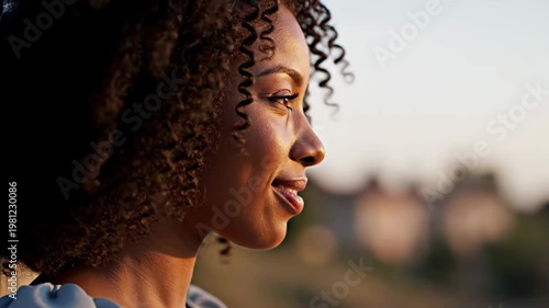 Serene profile portrait of a beautiful African American woman with curly hair, smiling in warm golden hour sunlight outdoors.