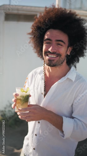 Happy young man with curly hair drinking a refreshing mojito cocktail while standing on a sandy beach