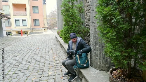 Man in Winter Clothes Resting on Stone Ledge in Cobblestone City Alley