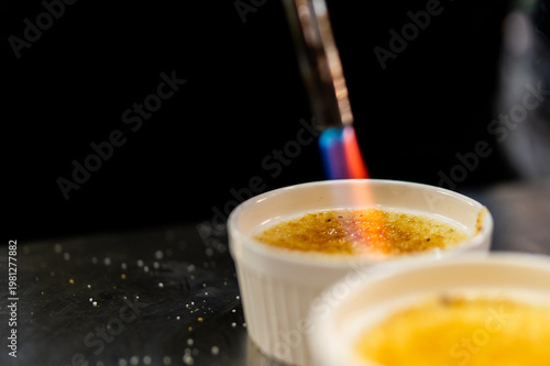 A kitchen torch carmelizing the sugar topping of a crème brûlée in a white ramekin, creating a golden-brown crust. Close-up shot with a dark background