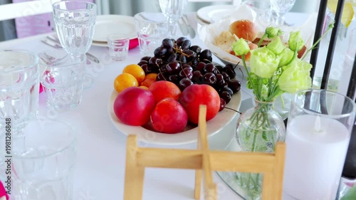 Pristine white plate displaying ripe peaches, grapes, apricots with elegant floral vase, presenting delicate dining composition highlighting seasonal fruit arrangement