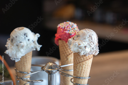 Three ice cream cones with different flavors and toppings on metal holder against cafe background, soft light and shallow depth of field. Food marketing, dessert advertising, cafe menu design