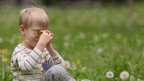 A fair-haired boy in a striped long sleeve sitting on green grass, holding a bright yellow dandelion flower to his eye. Close-up shot, blurred park background, daylight.
