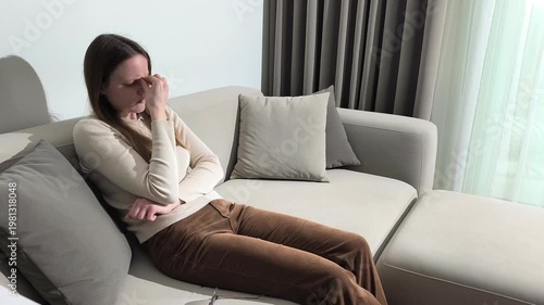 Woman massages nose bridge while sitting on sofa at home. Female presses face to relieve eye strain and head discomfort. Gesture reflects tension after screen use and need for short break..