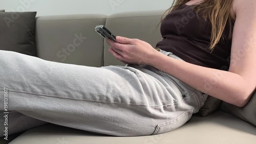 Woman sits on sofa holding smartphone and using mobile device at home. Female interacts with screen during rest. Concept digital lifestyle, communication, everyday routine, leisure time..