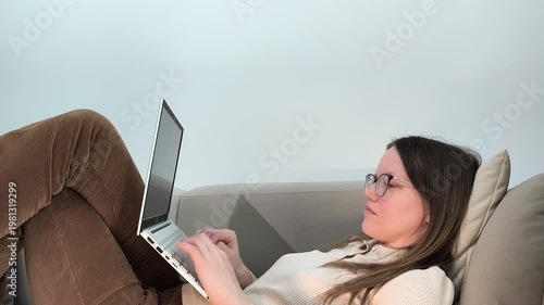 Laptop typing continues as woman lies on sofa in twisted pose working at home. Female uses computer during remote job in uncomfortable position. Lifestyle concept showing casual freelance