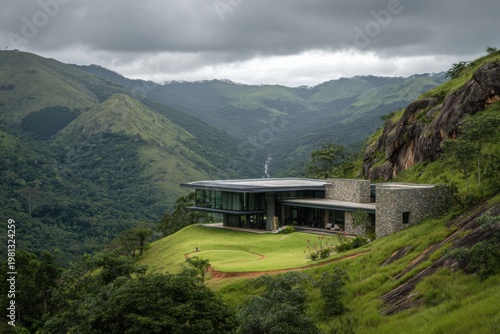 A contemporary house is nestled on a hillside, offering stunning views of lush green mountains beneath a cloudy sky, all set in a serene and isolated location