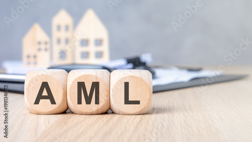 AML - Anti-Money Laundering - acronym on wooden blocks. Blurred background with miniature houses and magnifying glass. Real estate compliance concept.