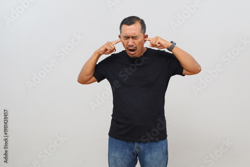 фотографія An adult man stands isolated on a white background, who is angry, covering his ears with his fingers and shouting