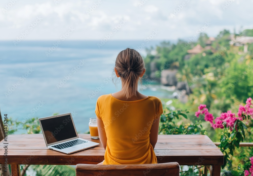 Fototapeta premium Woman sitting at a wooden table with a laptop and drink, enjoying remote work and freedom with a stunning ocean and green cliff view