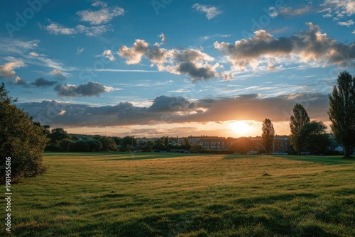 Grassy field leads to trees with sun setting behind distant buildings, cloudy sky