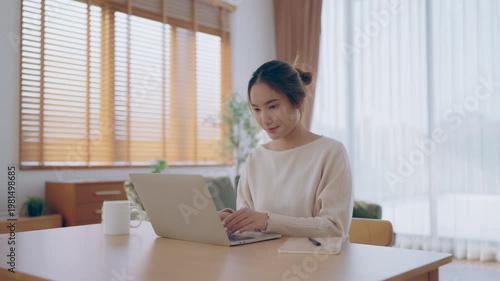 Young Asian woman using laptop computer for business studying, having video call with online virtual webinar training meeting, writing notes. Focused student female working in living room at home