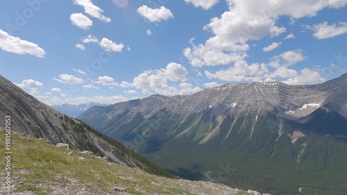 Scenic summer mountain landscape with dramatic rocky peaks, green forest valleys, and blue sky with clouds. View from a hiking trail overlooking the Canadian Rockies, capturing nature, adventure, and 