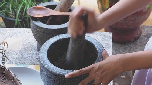 Authentic Thai cooking scene with fresh ingredients and traditional stone mortar and pestle. Hands preparing spicy sauce using chilies, garlic, lime, eggs, and herbs on a rustic kitchen table. Concept