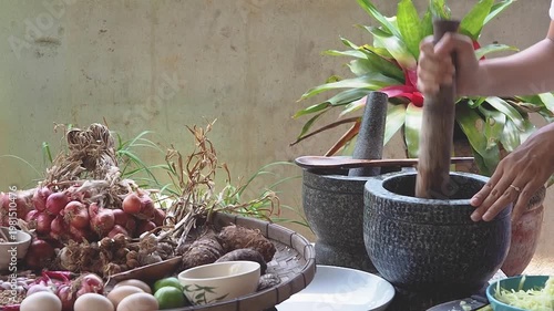 Authentic Thai cooking scene with fresh ingredients and traditional stone mortar and pestle. Hands preparing spicy sauce using chilies, garlic, lime, eggs, and herbs on a rustic kitchen table. Concept