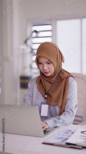 Asian adult woman working home office laptop computer typing reports analyzing data virtual meeting professional hijab attire focused remote business education technology