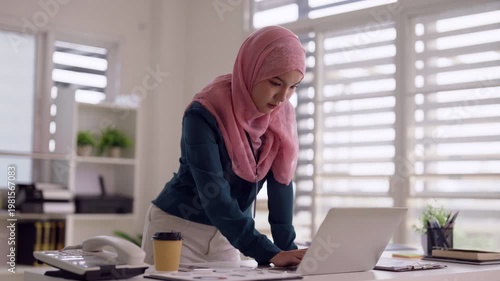 Adult Asian Muslim woman using laptop computer typing business reports working modern office focused remote work environment