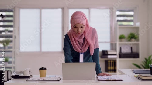 Muslim professional woman standing working on laptop computer analyzing business data report in a bright modern office environment