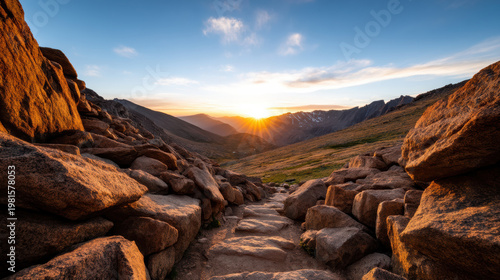 Rocky mountain trail at sunrise with sun rays peeking over distant peaks under a partly cloudy blue sky.