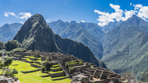 The famous Lost City of the Incas Machu Picchu. Dilapidated stone buildings, stairs, green terraces. Silhouettes of people walking. The Andes mountain range, Huayna Picchu, blue sky, clouds. Peru.
