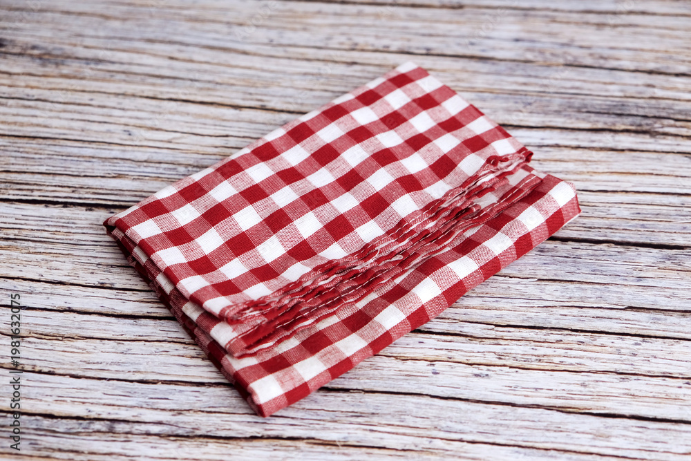 Obraz premium Red and white checkered cloth napkin rests on wood. Folds show soft texture and gentle creases. Rustic background adds charm and nostalgia. Ideal for kitchens, cafes, or picnic scenes