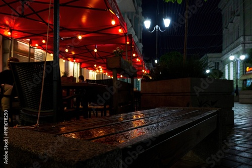 Rain wet wooden bench near outdoor cafe terrace with red canopy lights on cozy city street at night.