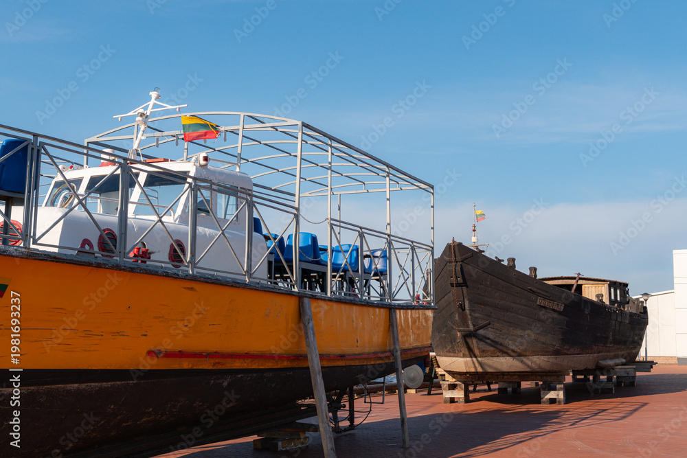 Fototapeta premium Bright yellow passenger ferry boat with metal cage seating structure rests beside a historic wooden black vessel upon a dry dock platform under a clear blue sky in summer daylight.