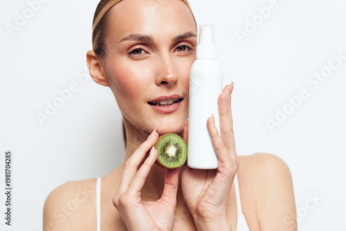 Woman with natural makeup holds cotton pad and kiwi slice near face, skincare routine concept, she touches cheeks, bright white background, beauty wellness self care fresh glow expression