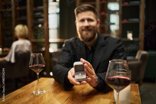 Photography Portrait of Caucasian young adult man presenting engagement ring box across restaurant table, proposing marriage