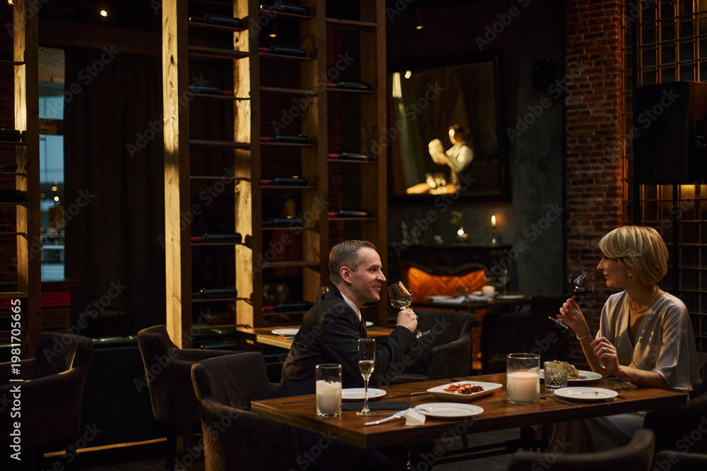 Fototapeta premium Caucasian middle aged couple dining and tasting wine in upscale restaurant, discussing during evening date. Ideal for luxury hospitality, dating, fine dining, lifestyle marketing