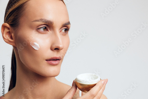 Young woman applying cream on face holding jar, skincare routine, natural beauty, clean skin, soft lighting, neutral background, focused expression, healthy lifestyle