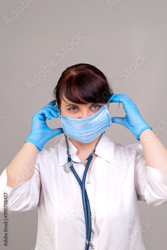 A doctor with a stethoscope in a white coat puts on a medical mask with blue gloves on a gray background