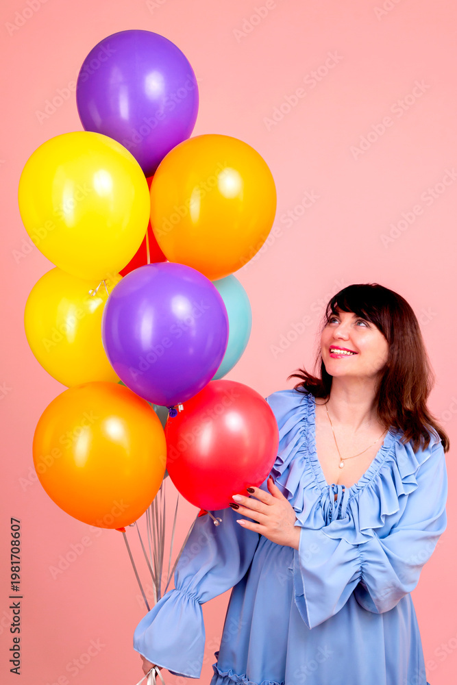 Obraz premium Happy young brunette woman in a blue dress with colorful balloons on a pink background