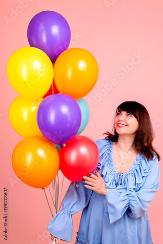 Happy young brunette woman in a blue dress with colorful balloons on a pink background