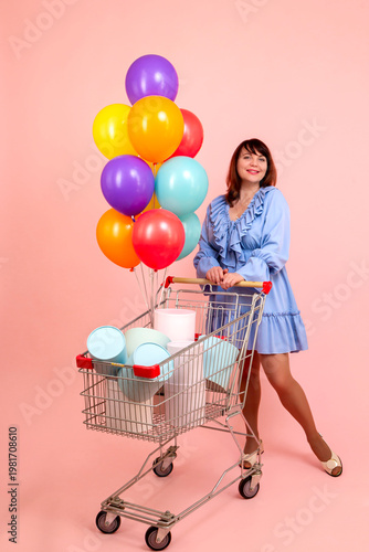 Brunette woman in blue dress with cart full of gifts and balloons on pink background