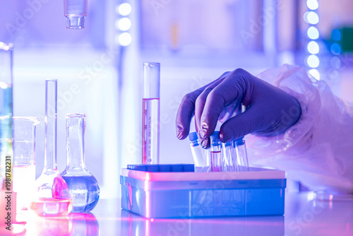 Gloved hand holding test tube in modern laboratory with sample vials and glassware under blue light, concept of research, biotech and clinical testing.