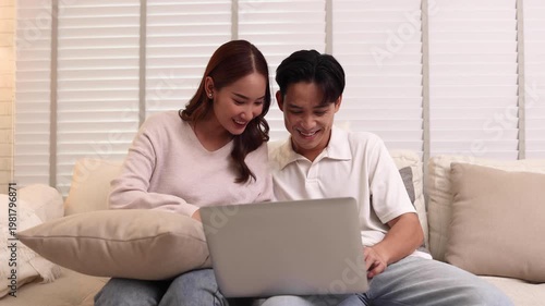 Happy young Asian couple smiles while sitting on a cozy white sofa, looking at a laptop screen together in their living room, perfect for home lifestyle, remote work, or online shopping concepts