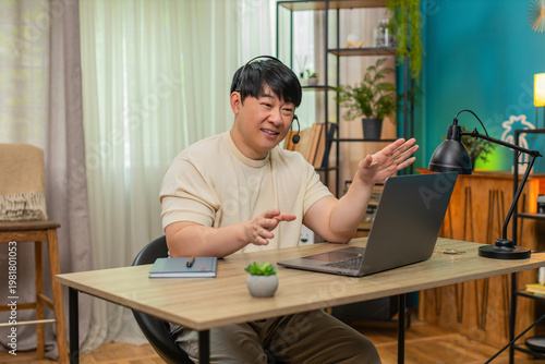Asian man sitting at home table speaks on laptop headset video call, providing customer support in professional tone. Chinese guy keeps serious posture, offering reliable help and clear guidance.