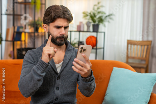 Indian man at home plays mobile casino on phone loses bet sighs holds head rubs forehead no gesture. Arabian guy on sofa sees result sad says no with hand dismayed mood after online gambling loss down