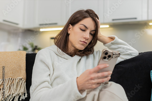 Young woman at home scrolls news on phone feels boredom fatigue stress low energy and low motivation. Caucasian girl on sofa swipes feed frowns sighs feels burnout mood and puts phone down for pause