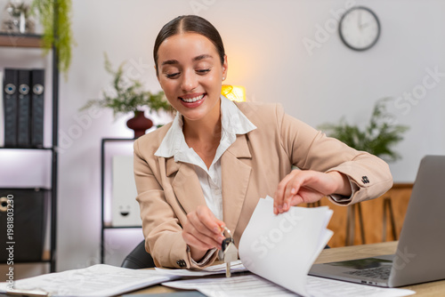 Young businesswoman at home office looks for keys in papers flips documents sighs and keeps searching. Freelancer girl at table finds keys smiles at own forgetful moment and returns to task on laptop